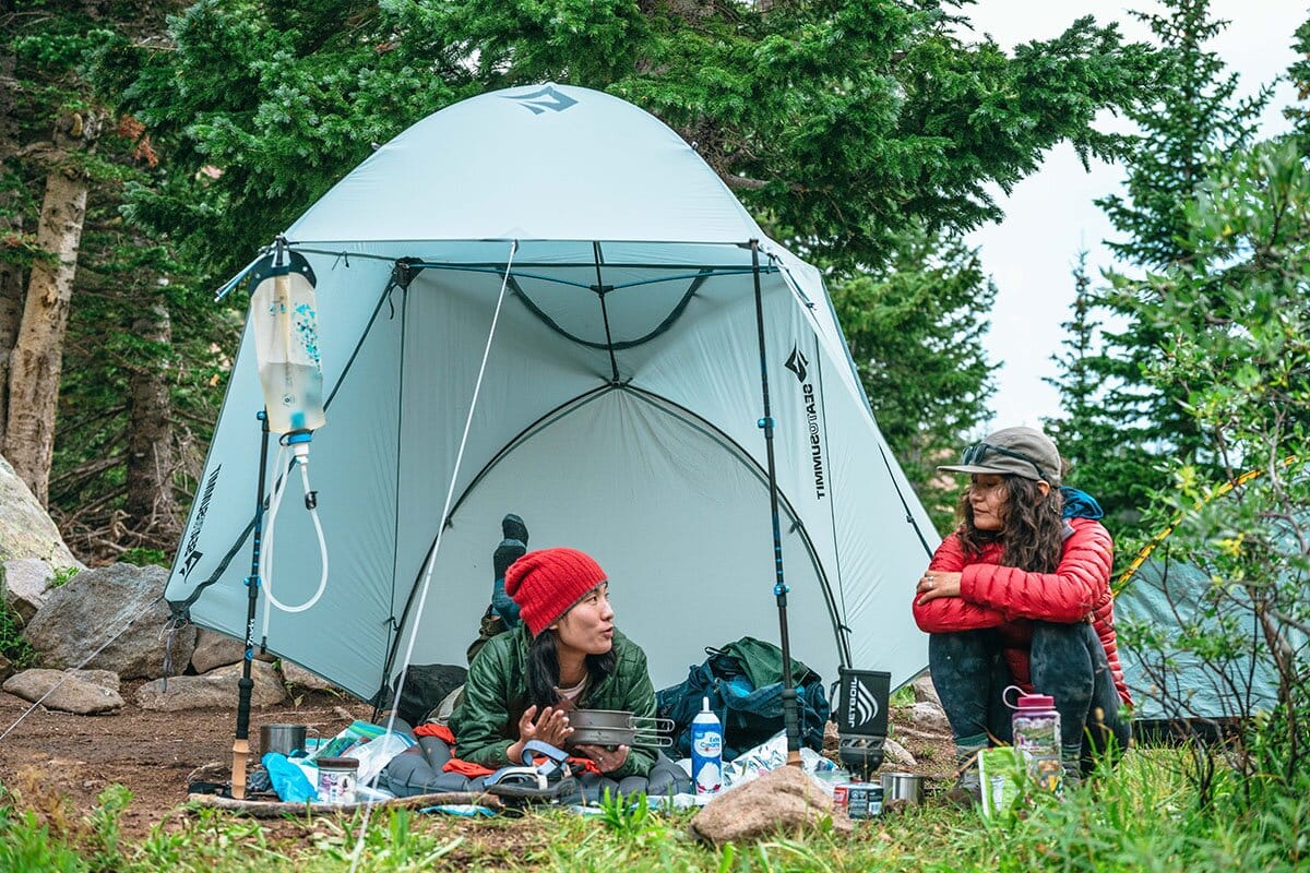 Two women lounging in front of Sea to Summit tent eating dinner and chatting mobile